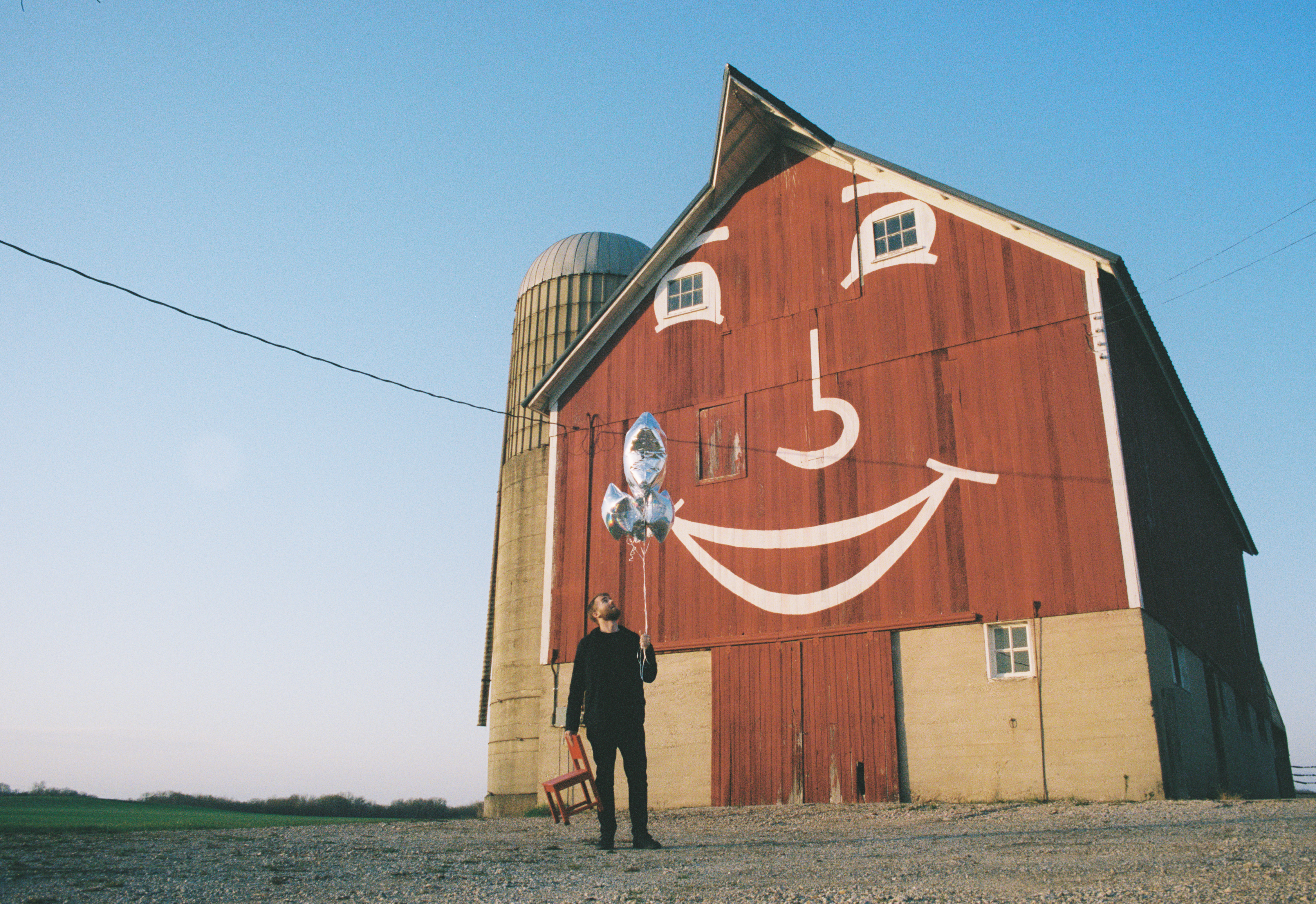 landscape photo of person, holding mylar balloons and a small red chair, in front of a red barn with a painted smiley face